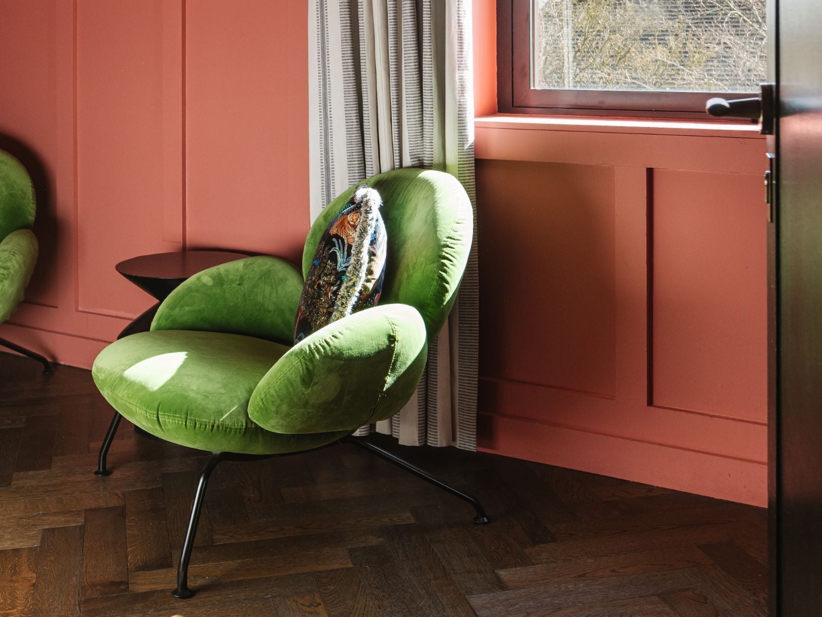 A green armchair with a patterned cushion, positioned next to a window with a view of trees.