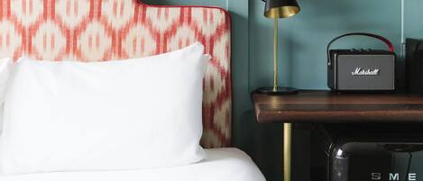 A hotel room with a patterned headboard, a bed with white linens, a Marshall speaker, a lamp, and a shelf with bottles and glasses.
