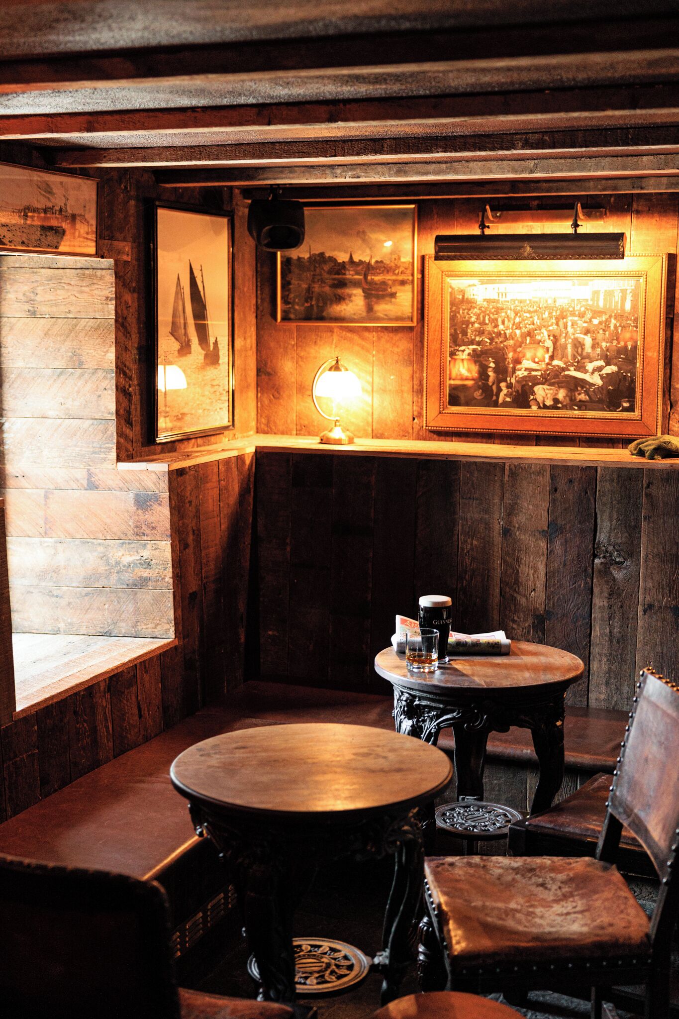 A cozy corner with wooden walls and a bench, featuring framed pictures and a table with a lamp.