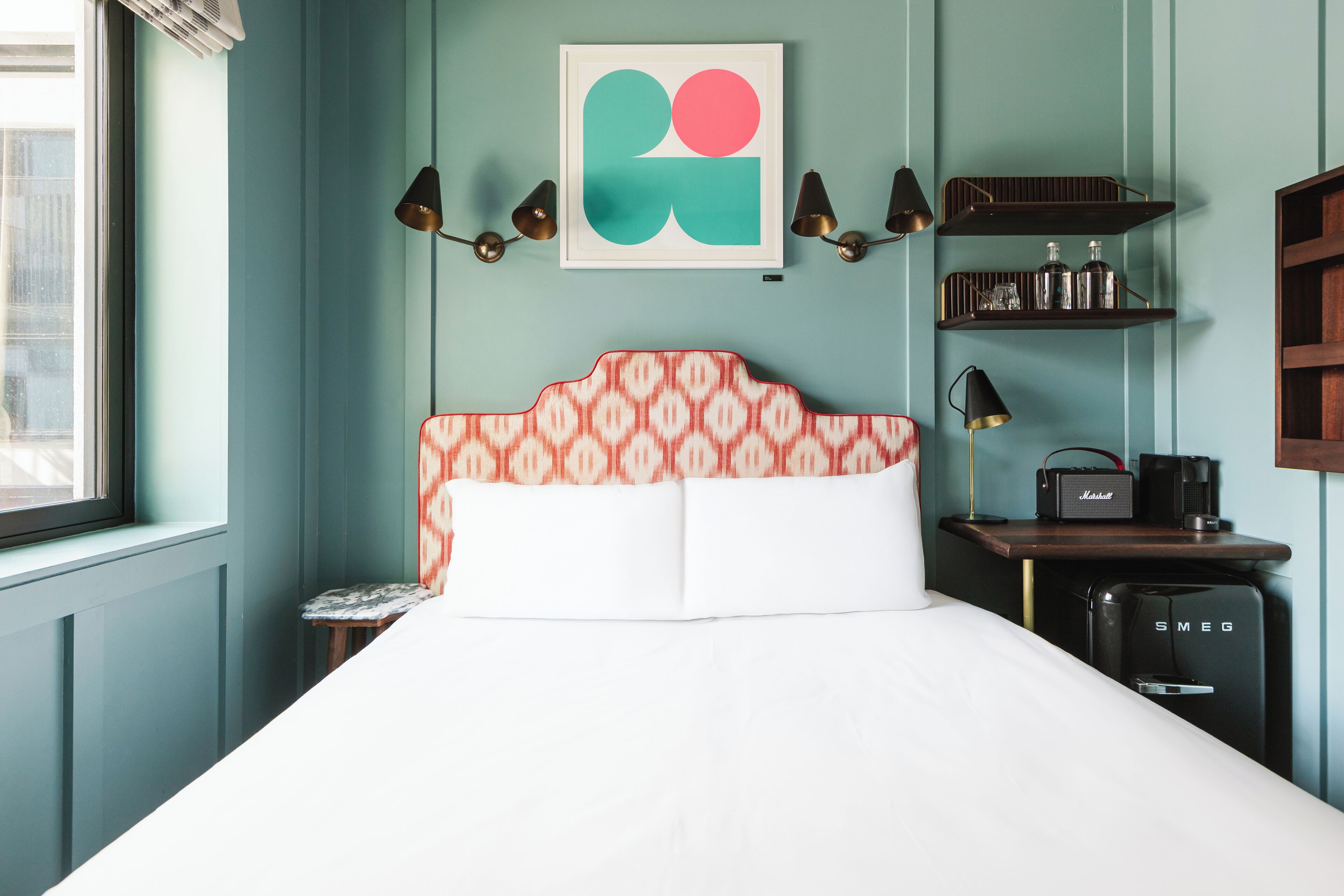 A neatly arranged bedroom with a bed, bedside table, lamp, and a small shelf with items.