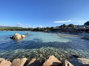 A coastal view with clear water, rocks, and a beach with buildings and trees in the background.