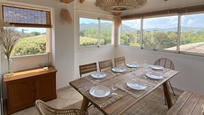 Un comedor con una mesa de madera preparada para seis personas, rodeado de sillas de mimbre, bajo un techo con vigas de madera y una lámpara de fibra trenzada.