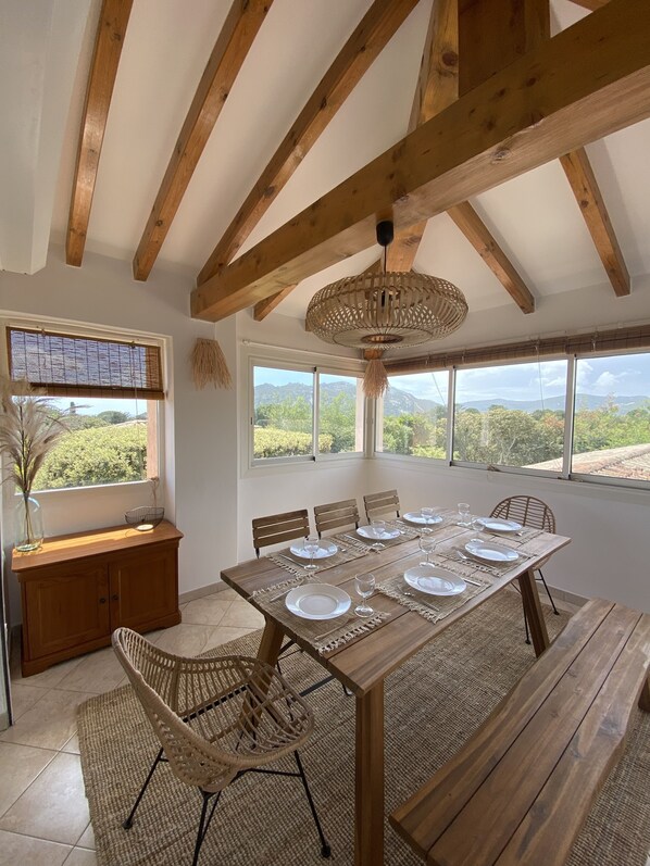 A dining area with a wooden table set for six, surrounded by wicker chairs, under a wooden beam ceiling with a woven light fixture.