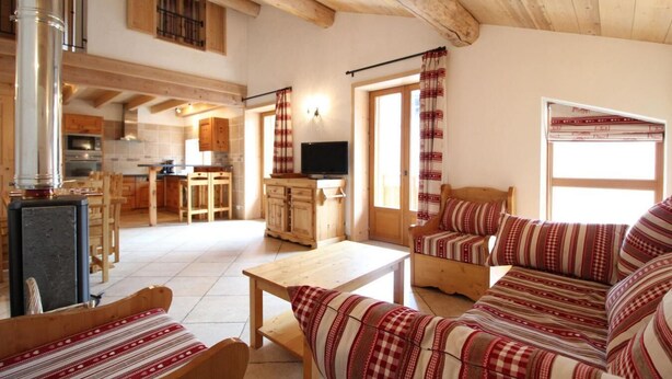 A cozy living room with a sofa, a wooden coffee table, and a television. There is a kitchen area with wooden cabinets and a window with curtains. - 100m from ski slopes, 1st floor, balcony, fireplace or stove, tv, ski locker, 90m², Val Cenis (Lanslebourg-Mont-Cenis)