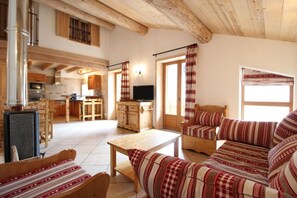 A cozy living room with a sofa, a wooden coffee table, and a television. There is a kitchen area with wooden cabinets and a window with curtains. - 100m from ski slopes, 1st floor, balcony, fireplace or stove, tv, ski locker, 90m², Val Cenis (Lanslebourg-Mont-Cenis)