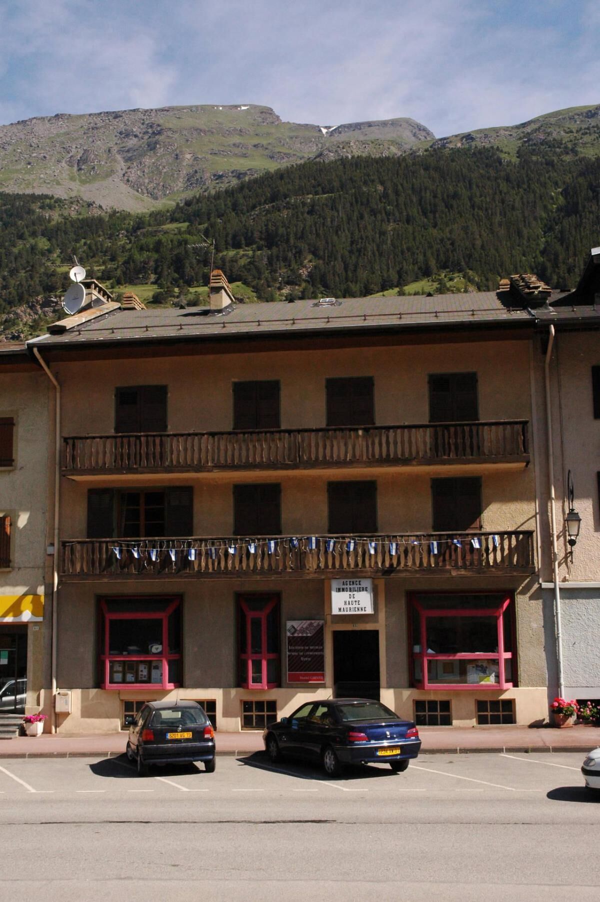 A two-story building with a balcony, a sign for an agency, and cars parked in front.