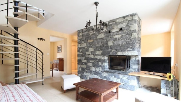 Una sala de estar con chimenea de piedra, televisor montado en una pared de piedra, mesa de centro de madera, sofá blanco y una escalera de caracol.