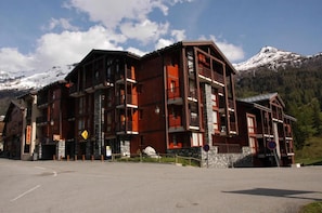 A multi-story hotel with balconies, a ski slope sign, and snow-capped mountains in the background.