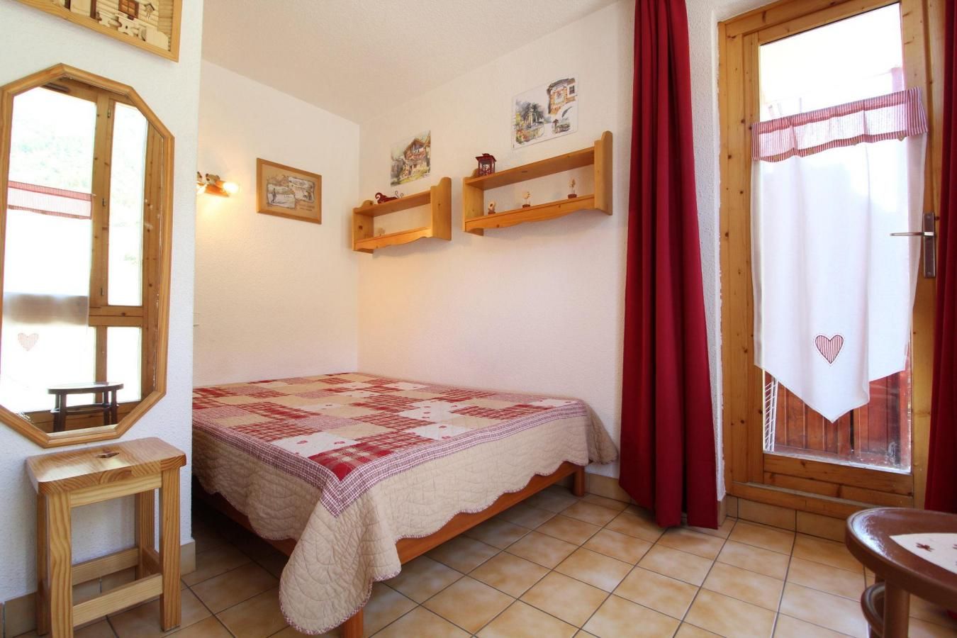 A bedroom with a bed, a wooden stool, a window with curtains, and wall-mounted shelves.