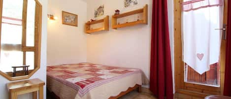 A bedroom with a bed, a wooden stool, a window with curtains, and wall-mounted shelves.