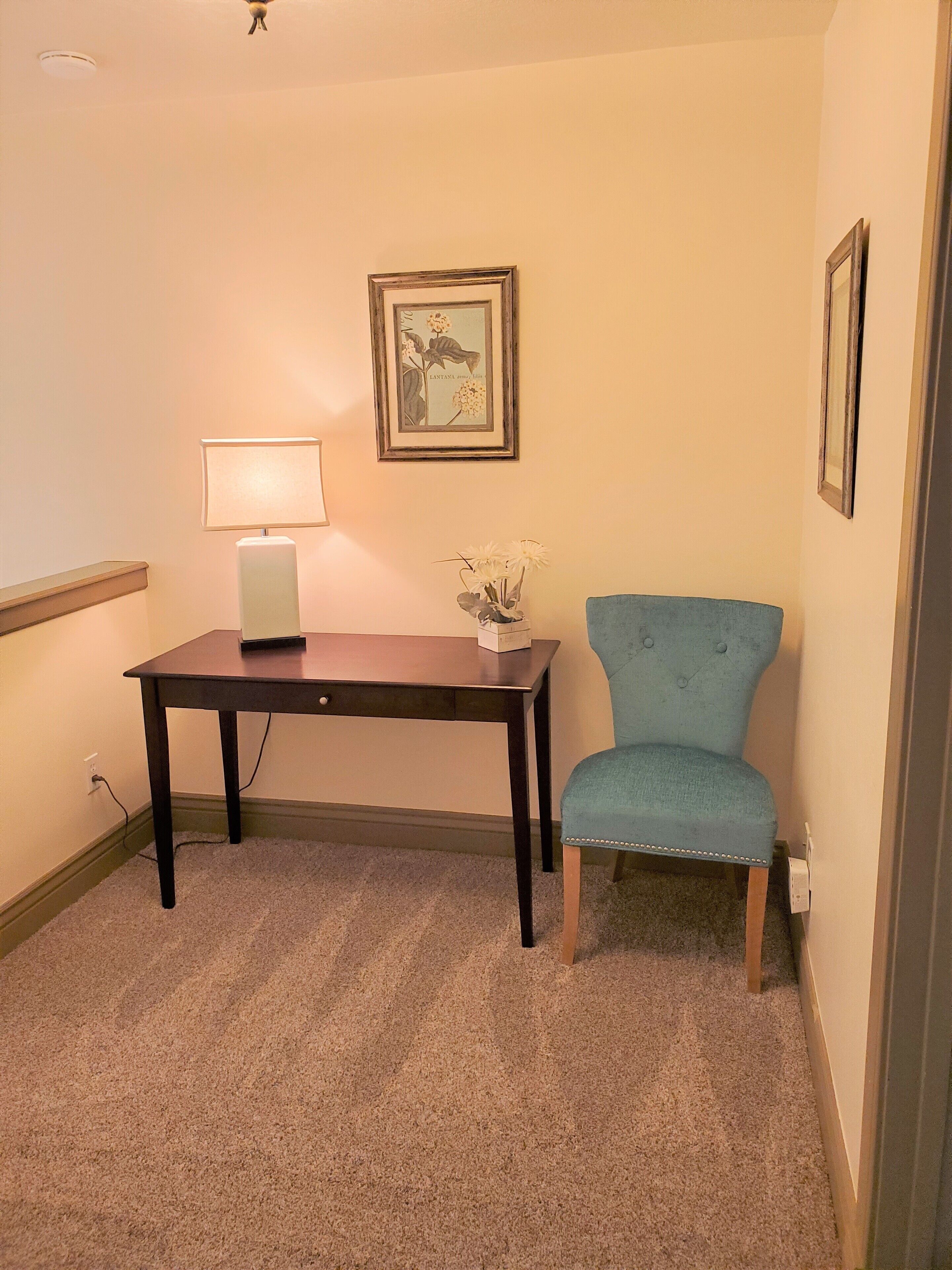 A small room with a dark wooden table, a blue upholstered chair, a wall-mounted lamp, and a framed botanical print.