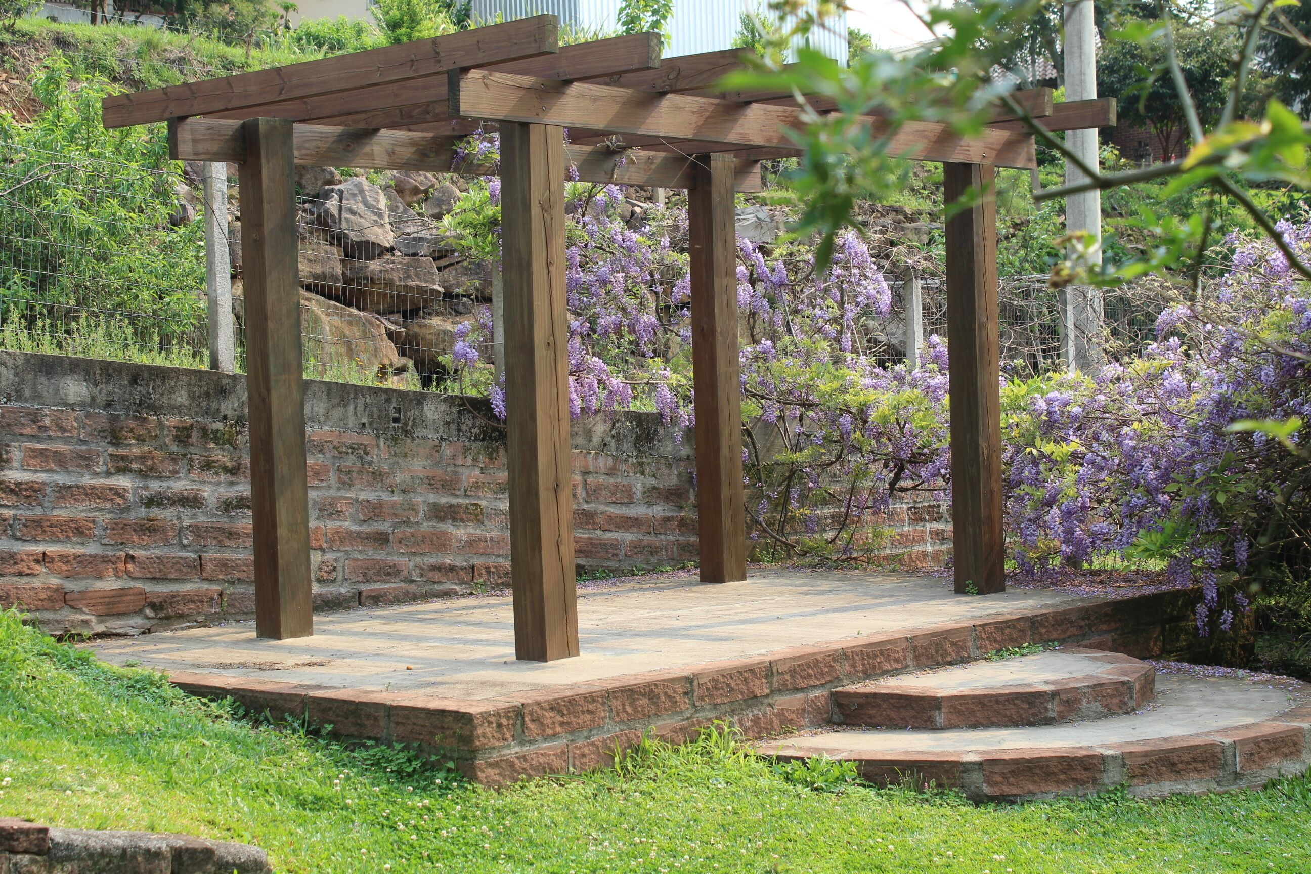 Eine Pergola aus Holz mit Steinstufen, umgeben von üppiger Bepflanzung und blühenden, violetten Pflanzen.