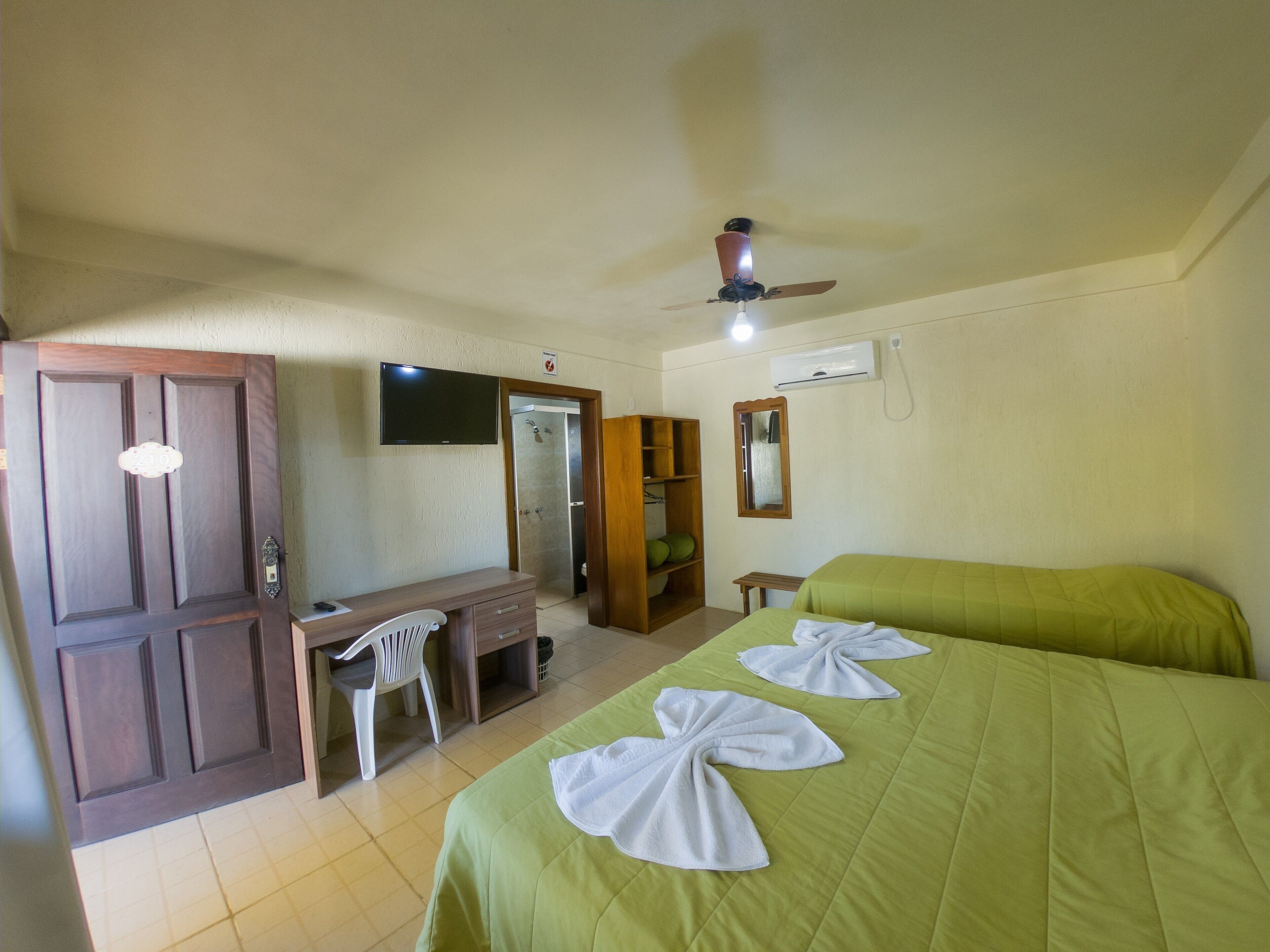 Una habitación de hotel con una cama, un escritorio, una silla, un armario, un espejo y un ventilador de techo.