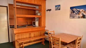Een compacte keuken met houten kastjes, een magnetron en een spoelbak. Er staat een houten bank en een tafel met stoelen. Twee ingelijste foto's hangen aan de muur.