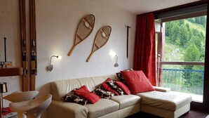 En hyggelig stue med en beige sofa, røde og mønstrede puder, et glasbord og udsigt til en balkon med gelænder.