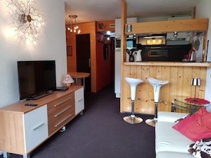 A compact hotel room with a TV, a desk, a chair, a bed, and a kitchenette.