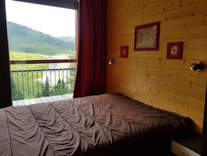 A bedroom with a bed, a wooden wall, a window with a view of a green landscape, and a balcony with a railing.