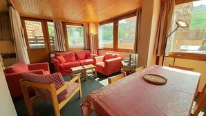 A cozy room with wooden ceiling, red sofas, a dining table with a tablecloth, and large windows with curtains.