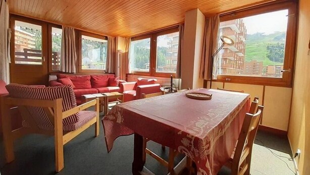 A cozy room with a wooden ceiling, a sofa set, a dining table, and large windows offering a view of buildings and greenery.