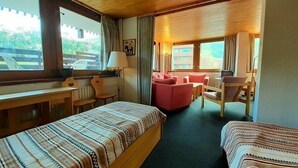 A room with two beds, a desk, a chair, a sofa, and a window with a view of greenery.
