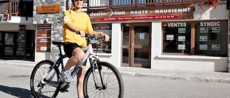 A person riding a bicycle in front of a building with a sign that reads "desinotion HAUTE - MAURIENNE" and "VENTES SYNDIC".