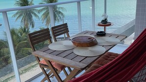 Ein Balkon mit Holztisch, Stühlen und einer Hängematte. Der Blick geht auf einen Strand und Palmen.