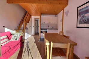 A cozy room with a wooden ceiling, a dining table, a pink sofa, and a staircase leading to a kitchen area.