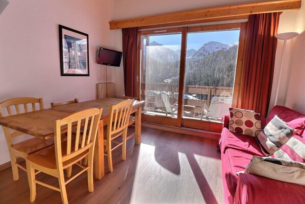 A room with a wooden dining table and chairs, a sofa with patterned cushions, a flat-screen TV, and a large window with a mountain view.