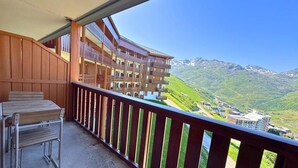 A balcony with a wooden railing, a table, and chairs. It overlooks a mountainous landscape with buildings in the distance.