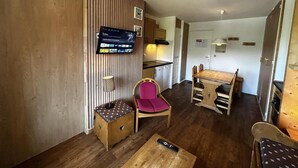 A compact living space with a wooden floor, a dining table with chairs, a small kitchen area, and a mounted flat-screen TV.