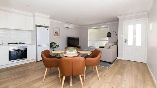 Een moderne keuken met witte kastjes, een eethoek met een ronde tafel en bruine stoelen, een bank en een televisie.