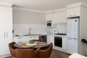 A modern kitchen with white cabinetry, a stainless steel sink, a microwave, a refrigerator, and a dining area with a round table and brown chairs. - Villa on Matthews - Modern & Cosy, Escape for 2 (Orange)