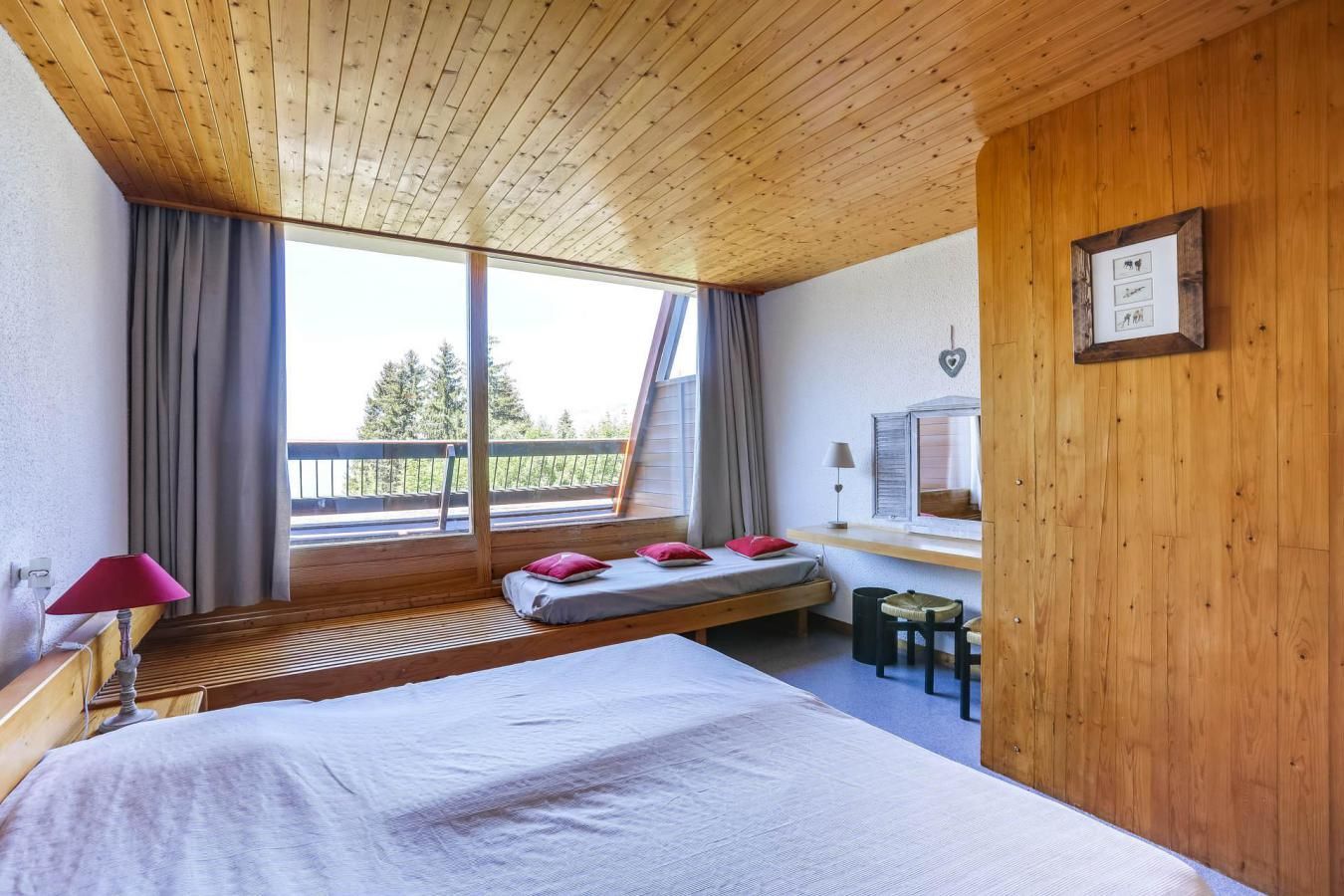 A bedroom with a large bed, a small sofa bed, a desk with a chair, and a window with a view of trees.