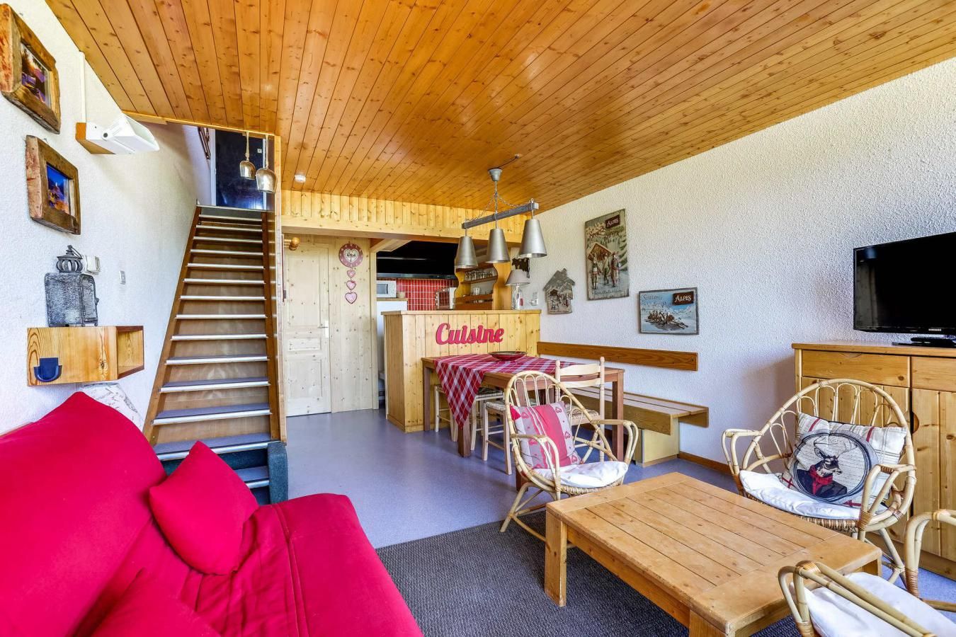 Una sala acogedora con un sofá rojo, una mesa de comedor de madera, sillas de mimbre, una zona de cocina con un letrero que dice 'Cuisine' y una escalera que conduce a otra habitación.