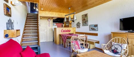 Una sala acogedora con un sofá rojo, una mesa de comedor de madera, sillas de mimbre, una zona de cocina con un letrero que dice 'Cuisine' y una escalera que conduce a otra habitación.
