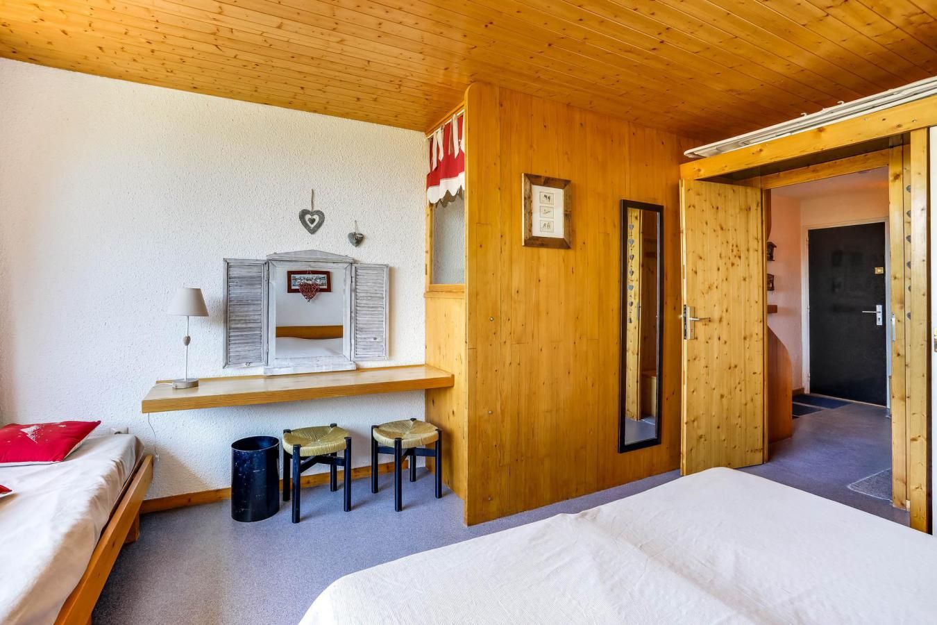 Una habitación con paneles de madera, una cama, un escritorio pequeño con una lámpara, dos sillas y una puerta que da a otra habitación.