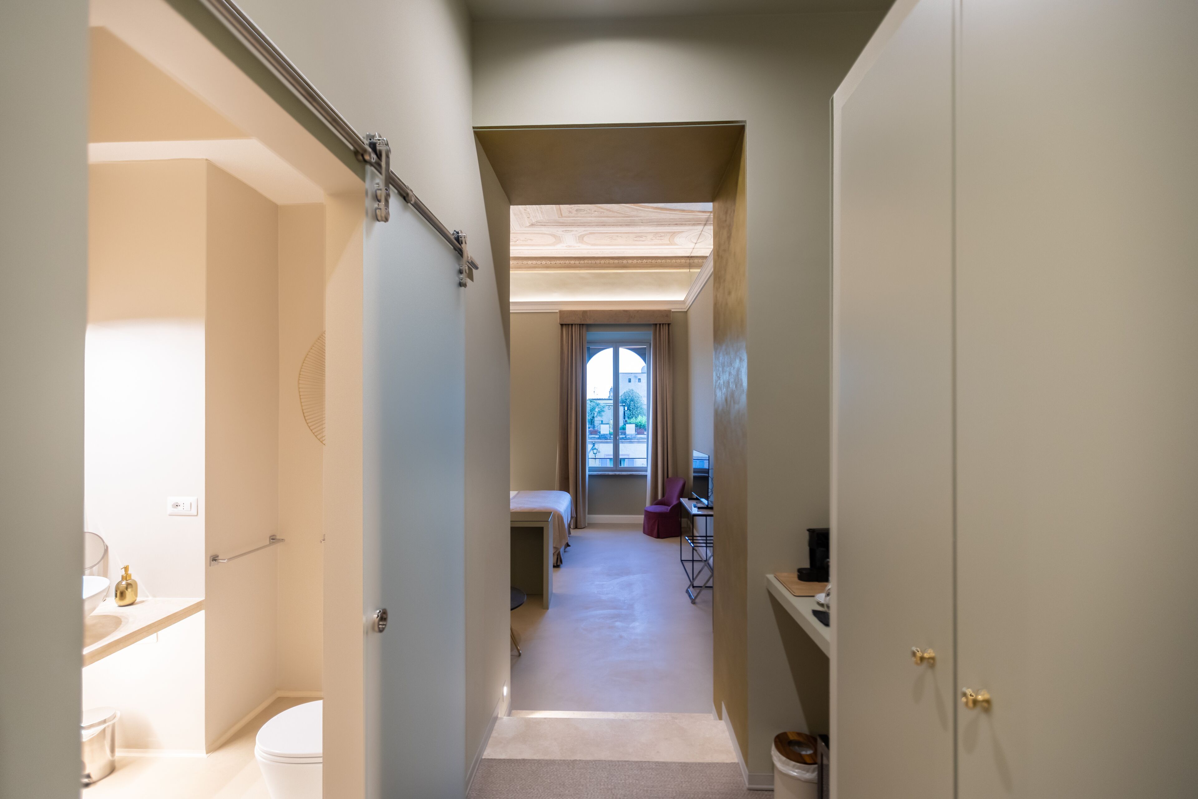 Un pasillo estrecho con un baño a la izquierda, una ventana con cortinas y un escritorio con una silla en el centro.