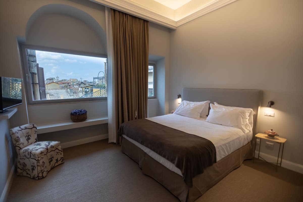 A hotel room with a large bed, a window with a view of buildings, a small table with a lamp, and a chair.