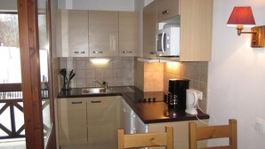 Cocina compacta con armarios de madera, microondas, cafetera y un pequeño comedor con dos sillas de madera.
