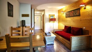 Una habitación acogedora con suelo de madera, un sofá de madera con cojines rojos, una mesa de comedor de madera con sillas y un televisor sobre un mueble de madera.