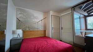 A bedroom with a wooden headboard, a red bedspread, a nightstand with a lamp, and a window with a view of trees. - 5th floor, balcony, tv, 56m², Brides Les Bains (Brides-les-Bains)