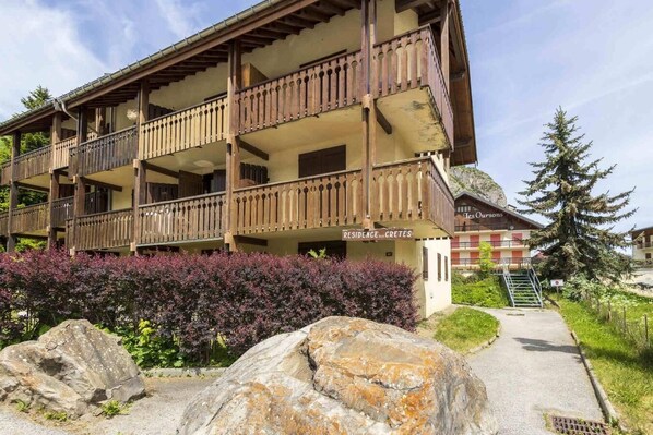 A multi-story building with balconies, a sign reading 'Residence Cretes,' and a surrounding garden with large rocks and shrubs.