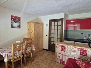 A compact room with a wooden dining table and chairs, a small kitchenette with red cabinets, and a door leading to another room. - 350m from ski lifts, 2nd floor, terrace, parking, tv, ski locker, 25m², Valloire (Valloire)