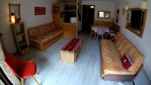 Ein kompakter Wohnbereich mit einer Couch, einem roten Sessel, einem kleinen Tisch und einer Küchenzeile.