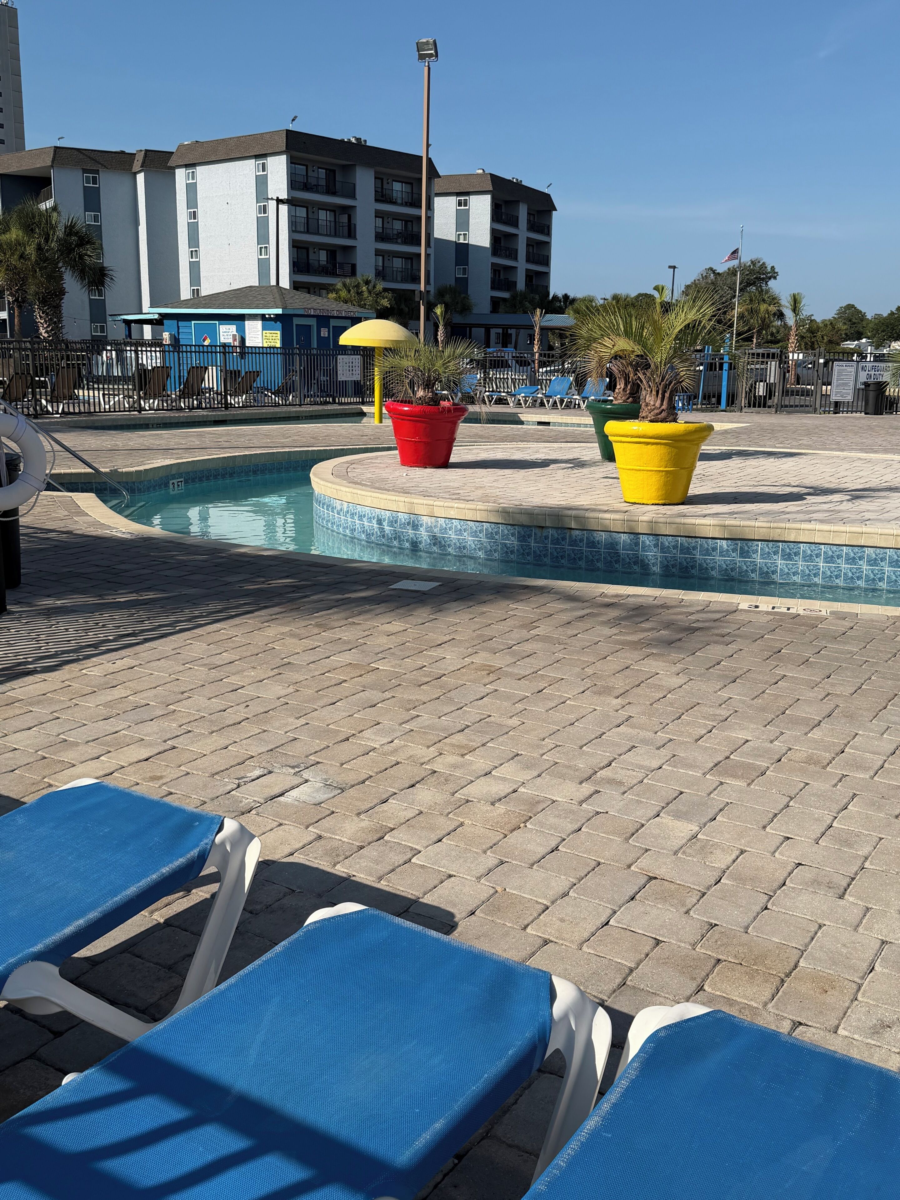 Zona de piscina amb gandules, vora de la piscina amb rajoles i un edifici al fons.