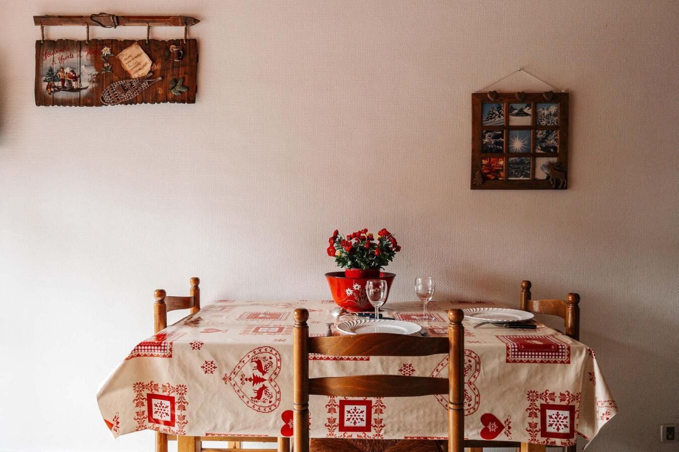 Une table à manger dressée avec une nappe, des chaises, un pot de fleurs rouge et un cadre photo fixé au mur.
