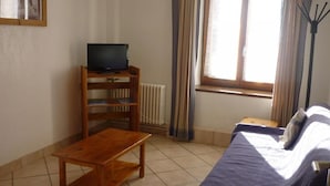 A small, well-lit room with a wooden table, a sofa, a television on a stand, and a window with curtains.