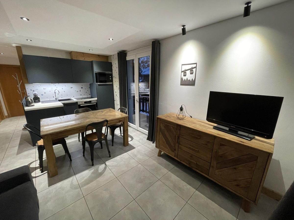 A modern kitchen with a dining area, a TV on a wooden cabinet, and a sliding glass door leading to a balcony.