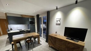 A modern kitchen with a dining area, a TV on a wooden cabinet, and a sliding glass door leading to a balcony.