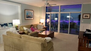 A living room with a beige sofa, a coffee table, and a TV stand. There is a ceiling fan and a window with blinds. - Luxury Penthouse 4BR Condo - Great for Families! (North Myrtle Beach)